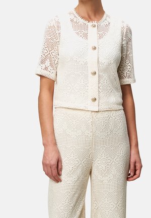 Mujer con una blusa blanca de ganchillo de manga corta con botones dorados y pantalones de ganchillo a juego de cintura alta.