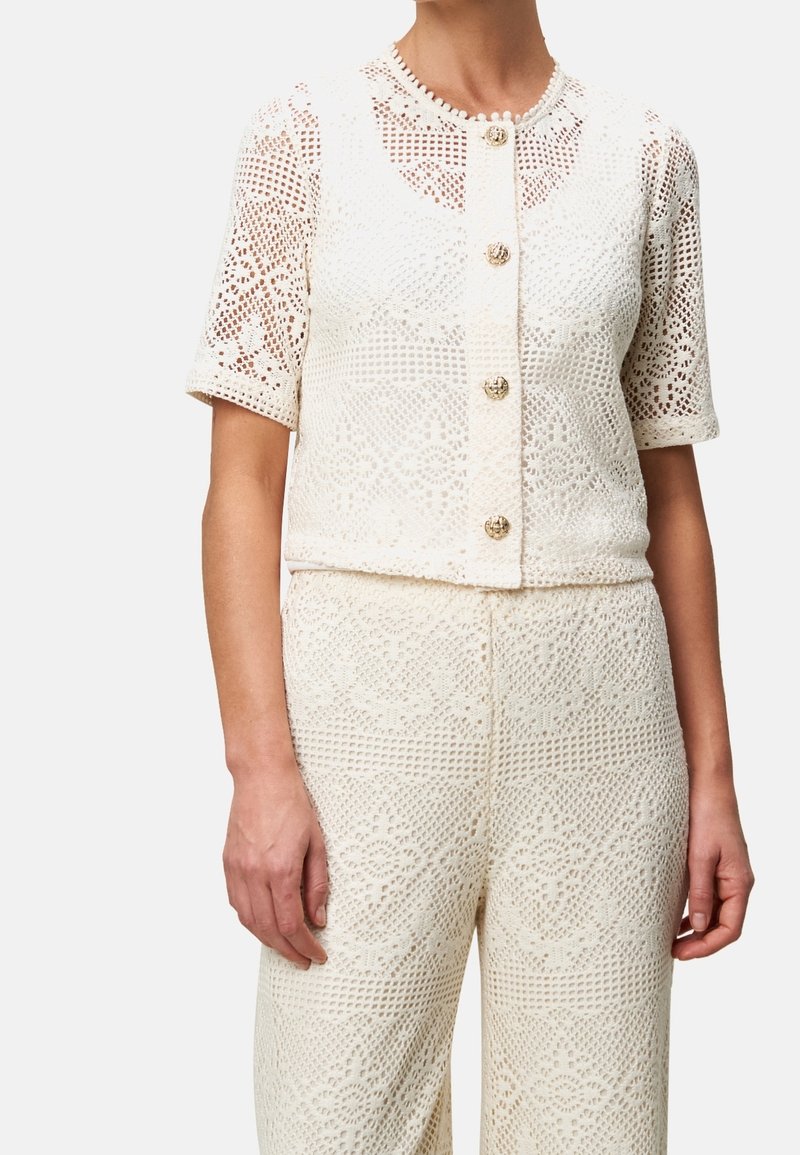 Femme portant un haut blanc en crochet à manches courtes avec des boutons dorés et un pantalon crocheté taille haute assorti.
