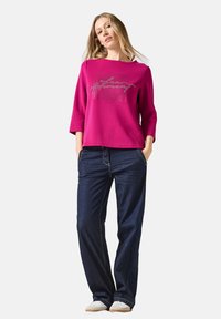 Sweatshirt fuchsia à manches trois-quarts, avec texte métallique et design graphique. Assortie avec un jean large bleu marine et des baskets blanches.