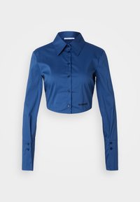 Przycięta niebieska koszula z długimi rękawami, zapinana na guziki z kołnierzykiem typu point i haftowanym logo przy dolnym brzegu. Gładki materiał i dopasowany krój.