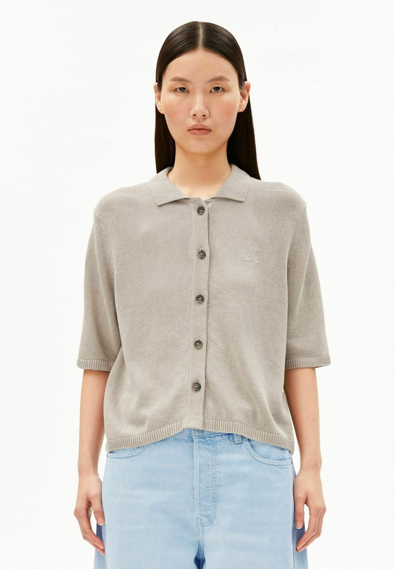 Woman with long dark hair wearing a beige button-up short sleeve sweater and light blue high-waisted jeans, standing against white background.