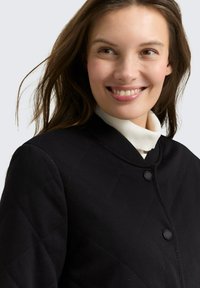 Czarna pikowana kurtka z okrągłym kołnierzem, z dwoma czarnymi guzikami i fakturowaną tkaniną. Noszona na kremowym golfie.