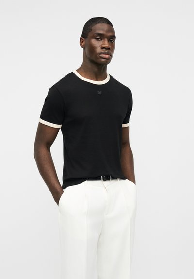 Άνδρας που φοράει μαύρο κοντομάνικο πουκάμισο με λευκή επένδυση και λευκό παντελόνι, στεκόμενος με τα χέρια στις τσέπες μπροστά σε απλό φόντο.