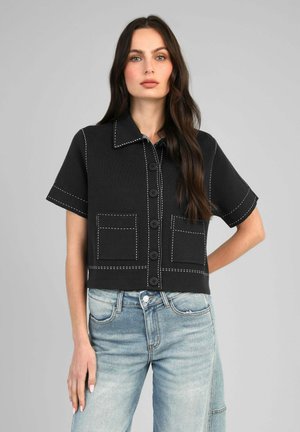 Femme aux longs cheveux foncés portant une chemise noire à manches courtes boutonnée avec des surpiqûres blanches et un jean bleu clair sur fond gris.