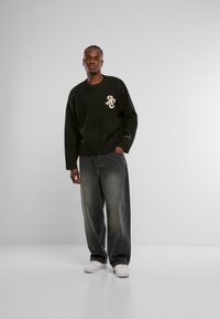 Suéter de cuello redondo verde oscuro con detalle de logotipo beige, combinado con jeans de mezclilla gris de pierna ancha y zapatillas blancas sobre un fondo liso.