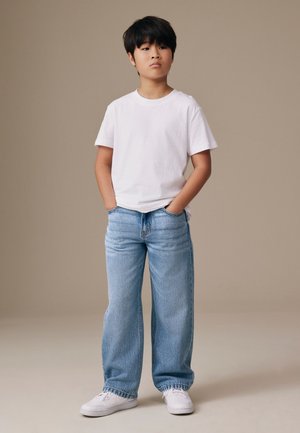 Fiatal fiú áll, kezét a zsebében tartja, fehér pólót, világoskék farmert és fehér sportcipőt visel, egyszínű bézs háttér előtt.