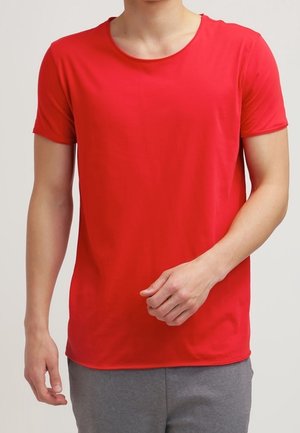 Mand iført en ensfarvet lys rød kortærmet T-shirt med rund hals og grå bukser, stående foran en hvid baggrund.