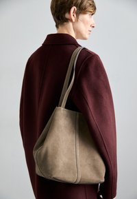 Tan suède hobo tas met een slouchy vorm en een contrasterende grijze schouderband, gepositioneerd naast een bordeauxrode jas. Afgeronde onderkant en stikseldetails.