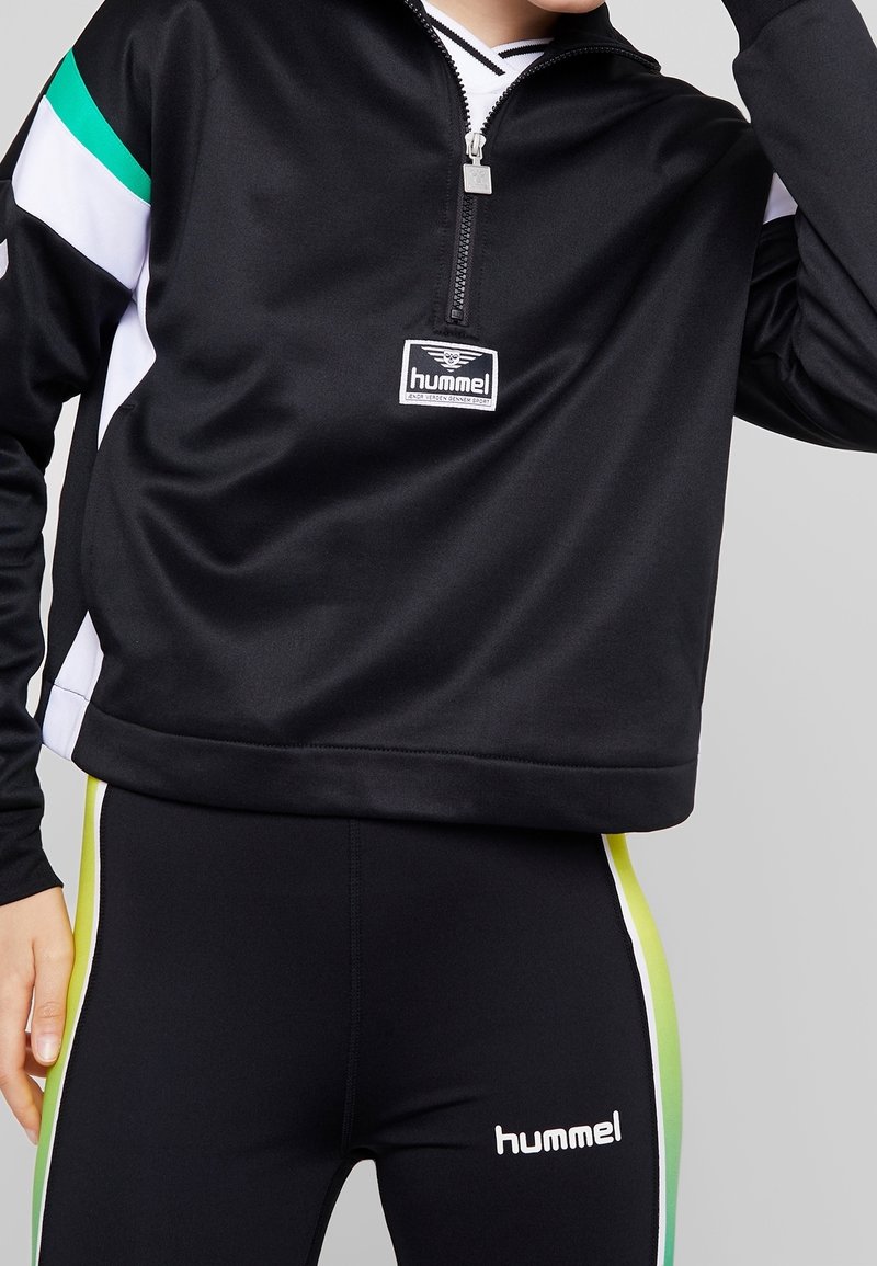 Personne portant une veste de sport noire Hummel avec zip et des leggings noirs avec des bandes latérales vertes et jaunes, se tenant devant un fond uni.