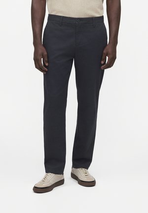 Mann trägt schwarze, gerade geschnittene Hose und beige Sneaker, beige gestricktes Oberteil teilweise sichtbar.