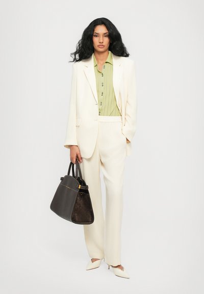 Weißer Blazer und Hose kombiniert mit einem grünen Hemd mit Knöpfen. Schwarze, strukturierte Handtasche mit goldenen Akzenten. Spitz zulaufende weiße High Heels.