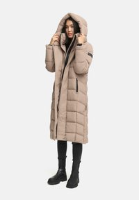 Beige lange pufferjas met een capuchon, met een gewatteerd ontwerp, zijritsas en een voorrits. G gedragen over zwarte broek en laarzen.
