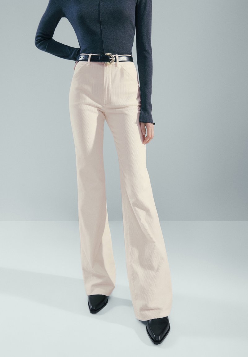 Pantalon évasé couleur crème en tissu texturé, associé à un haut bleu marine ajusté et une ceinture noire avec une boucle métallique. Chaussures noires pointues.