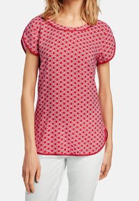 Haut rouge à manches courtes avec motif géométrique blanc, encolure ronde et bordure contrastante. Tissu doux ; coupe décontractée avec un ourlet arrondi.