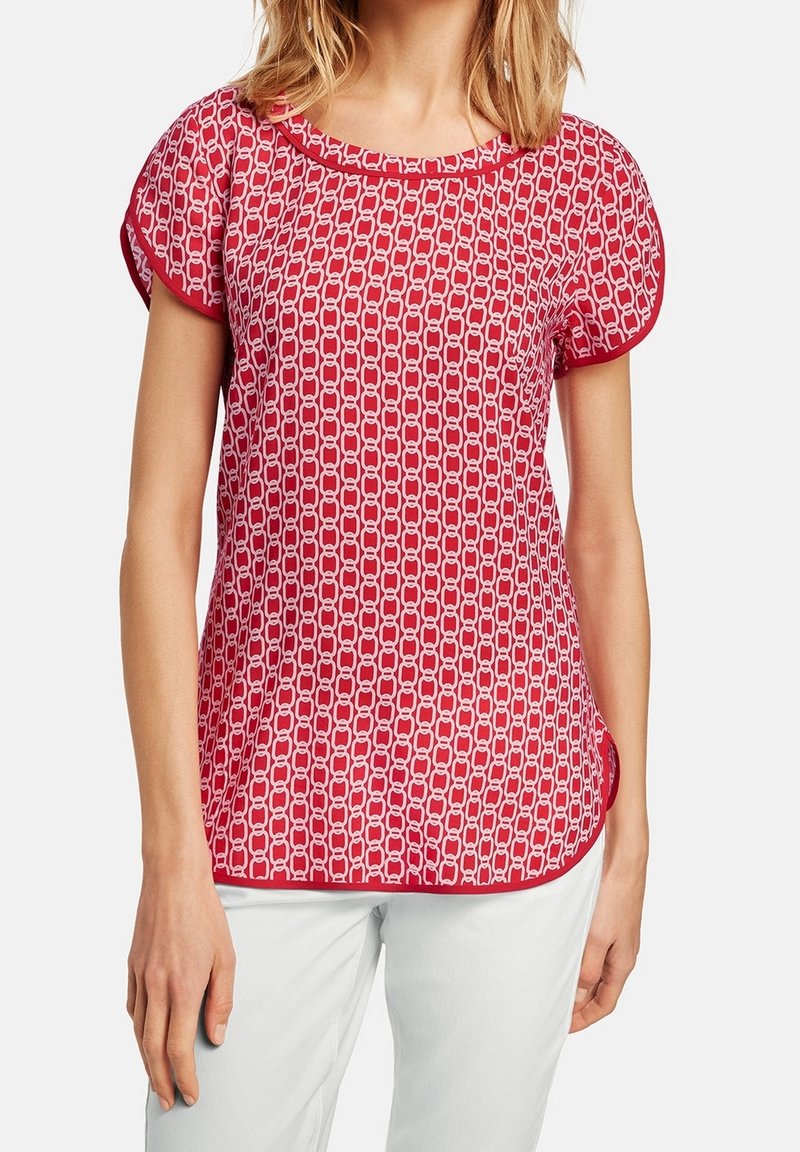 Haut rouge à manches courtes avec motif géométrique blanc, encolure ronde et bordure contrastante. Tissu doux ; coupe décontractée avec un ourlet arrondi.