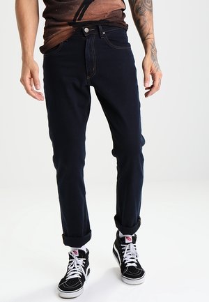 Homme portant un jean bleu foncé retroussé, des baskets montantes noires avec des lacets blancs et une chemise marron, debout sur un fond blanc.