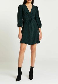 Vestido de estampado animal verde con un escote en V profundo, detalle de cintura anudada y mangas abullonadas; combinado con botines negros hasta el tobillo. Textura de tela suave.