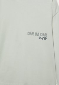 Light green sweatshirt featuring grey text "DAN DA DAN" and Japanese characters, made of soft fabric with a smooth texture.