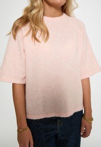 Gaiši rozā, pūkaina džemperis, kas izgatavots no mīksta auduma ar īsām piedurknēm un noapaļotu apkakli. Tam ir nedaudz brīvs piegriezums un negludā apakšmala.