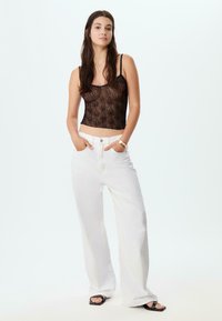 Czarny koronkowy crop top z cienkimi ramiączkami, w połączeniu z luźnymi, białymi spodniami typu wide-leg. Modelka pozująca z dłońmi w kieszeniach, w czarnych sandałach.
