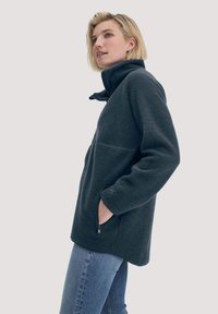 Giacca in fleece grigio scuro con colletto alto, tasche con zip e maniche lunghe. Texture morbida, vestibilità rilassata e dettagli delle cuciture discreti.