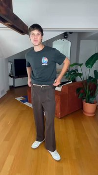 T-shirt preta com um gráfico, calças largas castanhas e ténis brancos. Ambiente interior com chão de madeira e plantas de interior visíveis.