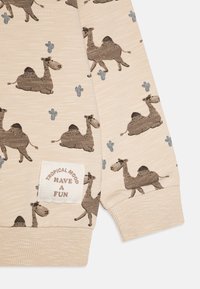 Camicia beige a maniche lunghe con motivo di cammelli marroni e cactus blu. Include una patch bianca con il testo "TROPICAL MOOD HAVE A FUN".