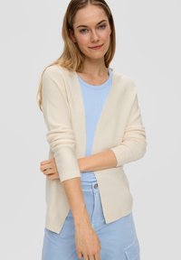 Romige cardigan met een ontspannen pasvorm, lange mouwen en een V-hals. Gedragen over een lichtblauw shirt met een gestructureerd breipatroon.
