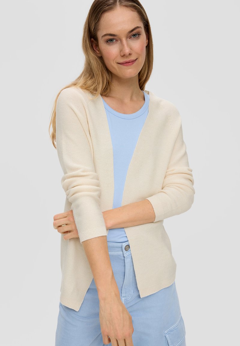 Romige cardigan met een ontspannen pasvorm, lange mouwen en een V-hals. Gedragen over een lichtblauw shirt met een gestructureerd breipatroon.