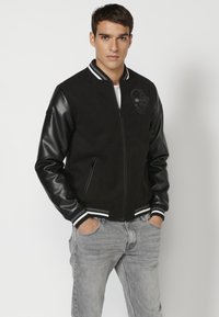Black bomber jacket with leather sleeves, striped ribbed cuffs and collar, front zipper, and skull graphic detail on the chest. Gray jeans worn.