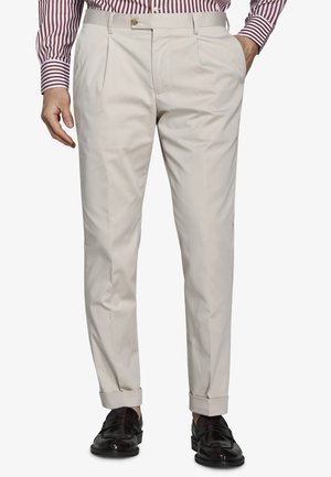 Mężczyzna w beżowych plisowanych spodniach z mankietami przy kostkach, w połączeniu z paskowaną koszulą i czarnymi skórzanymi mokasynami.