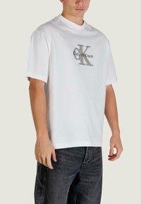 Biała bawełniana koszulka z okrągłym dekoltem, ozdobiona srebrnym logo z napisem "Calvin Klein" i "CK". Krótkie rękawy, luźny krój, minimalistyczny styl.