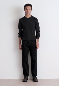 Maglietta nera a maniche lunghe, pantaloni neri a gamba dritta e scarpe marroni. Il tessuto appare solido e liscio senza motivi o dettagli visibili.