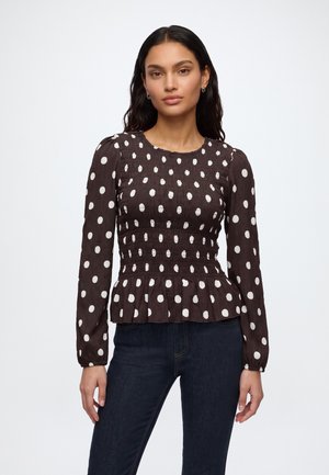 Femme aux longs cheveux foncés portant un chemisier noir ajusté à manches longues avec des pois blancs et un jean bleu foncé, debout sur un fond blanc.