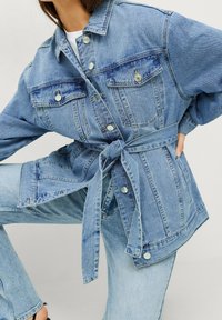 Jeansjacka i ljusblått med långa ärmar, två bröstfickor och knytning i midjan. Har metallknappar och en strukturerad design.