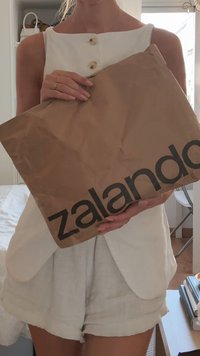 Brown crumpled paper bag with black "zalando" logo, clasped in hands. Background features white furniture and a light-colored bedspread.