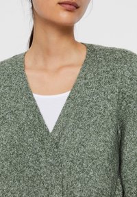 Green knitted cardigan with a V-neck design, featuring a soft, textured finish. Layered over a white shirt, highlighting versatility.