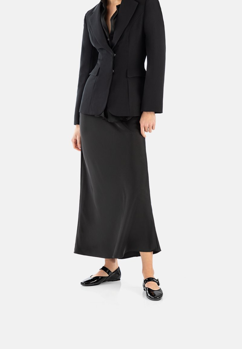 Blazer noir ajusté avec un col cranté, associé à une longue jupe en satin noir et des chaussures Mary Jane noires brillantes.