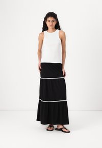 Débardeur blanc à côtes associé à une jupe maxi noire à volants avec une bordure blanche. Le mannequin porte des tongs noires. Tenue simple et polyvalente.