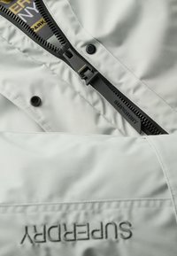 Jasnoszara kurtka ocieplana z czarnym zamkiem i logo. Posiada napy oraz gładką fakturę; czysty design z minimalnym wykończeniem.