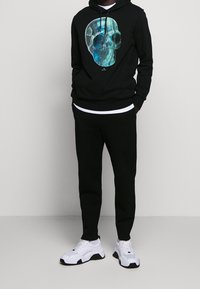 Černá mikina s kapucí s barevným motivem lebky, doplněná černými kalhotami a bílými teniskami s černými detaily. Ležérní a pohodlný střih.