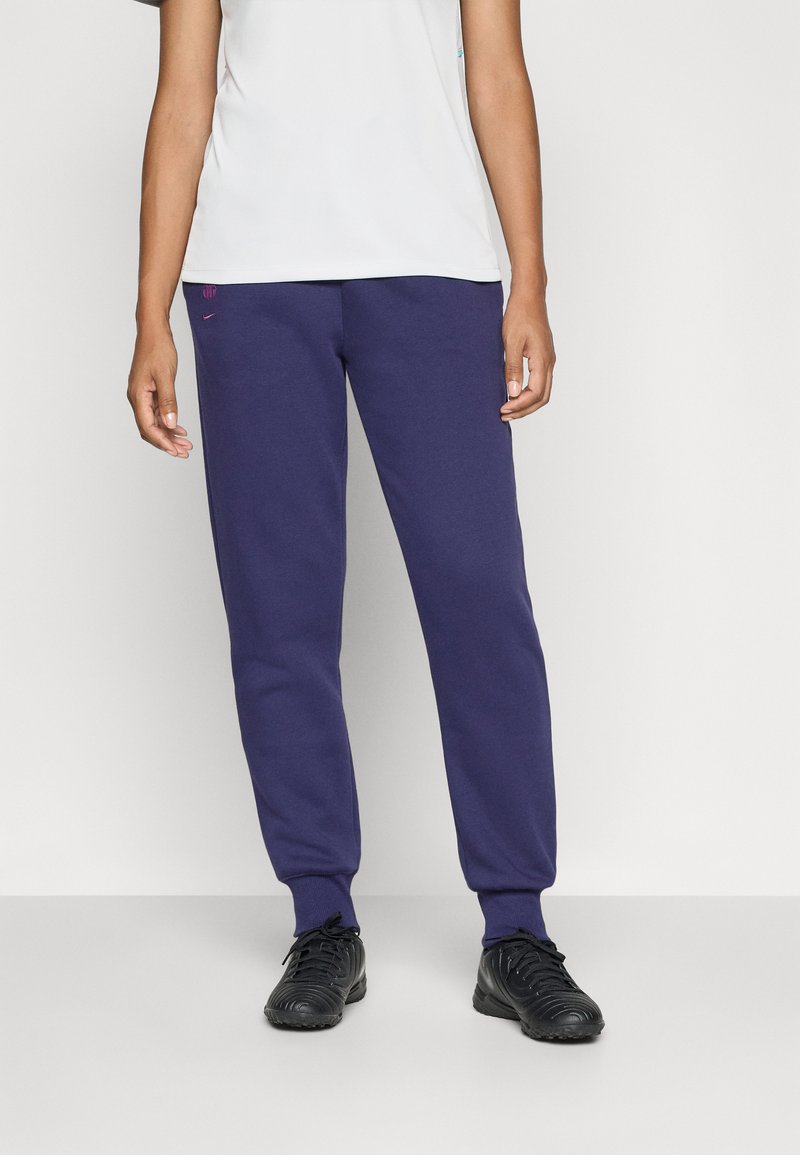Persona vestida con pantalones jogger azul marino y zapatillas deportivas negras, de pie contra un fondo gris claro liso.