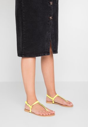 Jalat, joissa on keltaiset remmijalkineet ja musta nappihame, jossa on edessä halkio, seisomassa sileällä vaaleanharmaalla lattialla.