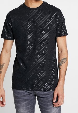 Czarny t-shirt z krótkim rękawem z błyszczącym, ukośnym napisem z nazwami miast Paryż, Londyn, Mediolan oraz cyframi rzymskimi, noszony przez osobę z tatuażami.