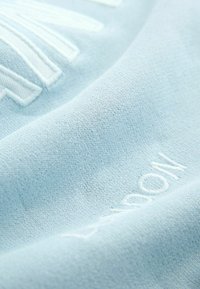 Tessuto azzurro chiaro con una trama testurizzata, caratterizzato da lettere ricamate bianche che accentuano la superficie, creando un sottile contrasto.