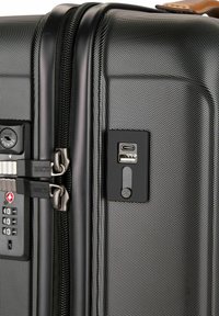 Valigia rigida nera con superficie textured. Dotata di doppie cerniere, lucchetto combinato, porte USB e una maniglia in pelle marrone.