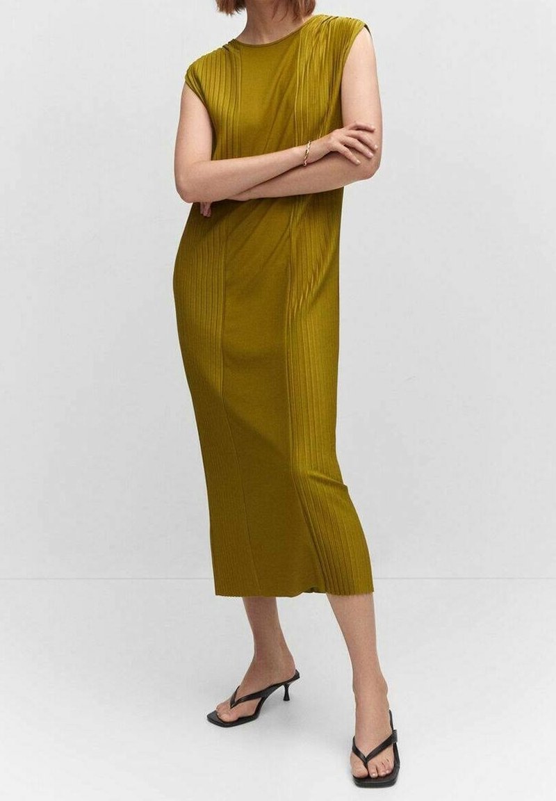 Robe vert olive à manches courtes, texture côtelée, longueur au genou. Portée avec des tongs noires, bras croisés dans un cadre neutre.