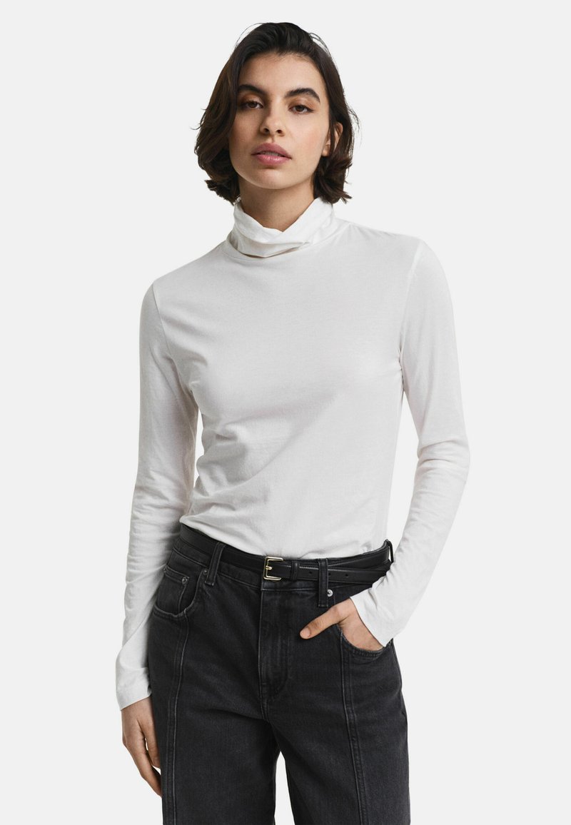 Vrouw in een witte lange mouw coltrui, gestopt in hoge zwarte jeans met een zwarte riem, met één hand in haar zak.