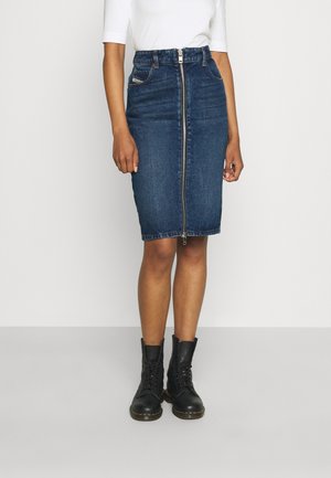 Gonna a tubino in denim blu scuro, con chiusura frontale a zip e tasche laterali. Indossata con stivali neri con lacci.