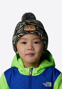 Czapka dziana z czarno-białym wzorem zebry, z pomponem i kolorową naszywką z tygryskiem. Widoczna jasna zielona i niebieska kurtka pod spodem.